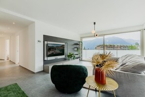 A modern living room with a flat-screen TV, a dark green chair, a round gold table with a vase, and a grey sofa.