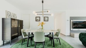 A modern dining area with a round table, six chairs, a green rug, a chandelier, and artwork on the walls.
