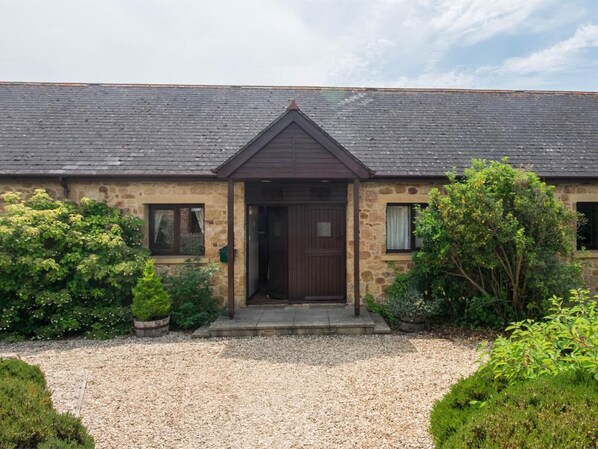 A stone building with a dark brown door, a gravel driveway, and surrounding greenery. - 2 Bed in Bridport (oc-dc162) (Chideock)