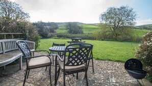 Ein Außenbereich mit Sitzgelegenheiten, einem Tisch, Stühlen und einem Grill, mit Blick auf eine grüne Landschaft.