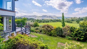 Une maison moderne dotée d’une grande baie vitrée, d’une terrasse en bois et d’un jardin bien entretenu, agrémenté de diverses plantes et offrant une vue sur des collines ondoyantes.