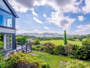 Ein modernes Haus mit einem großen Panoramafenster, einer Holzterrasse und einem gepflegten Garten mit verschiedenen Pflanzen und Blick auf sanfte Hügel.