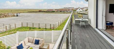 A balcony with outdoor furniture, a view of the beach, and a building in the background.