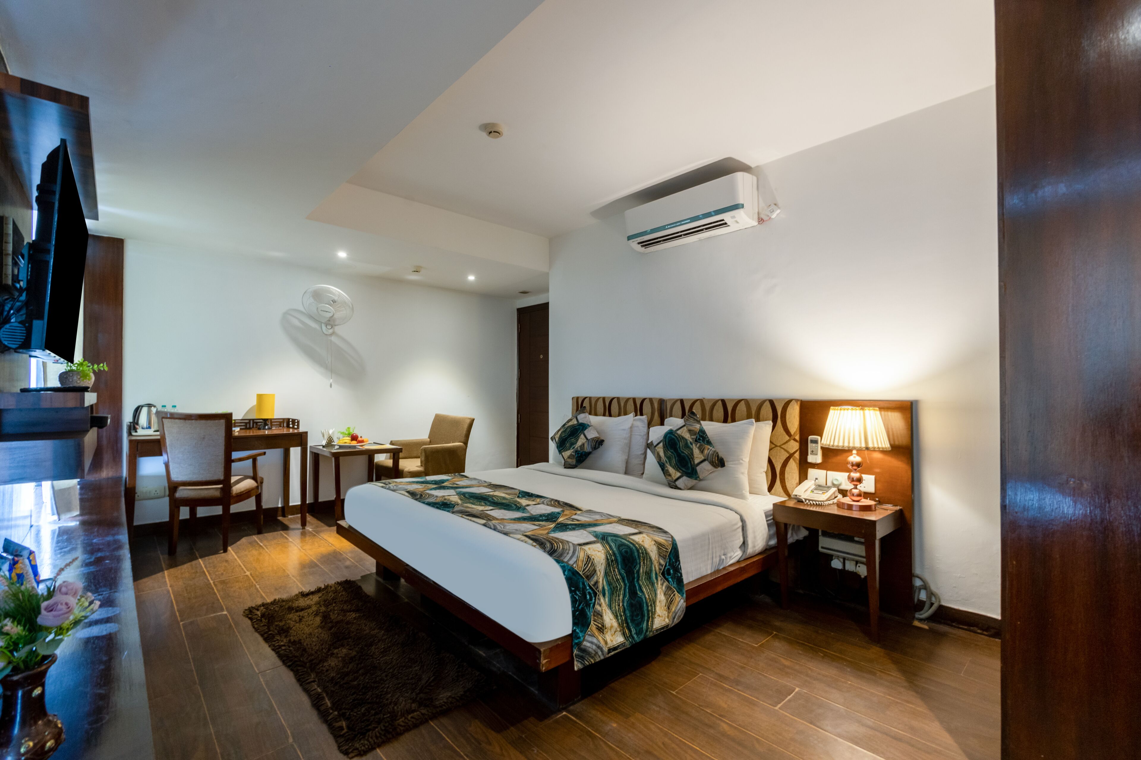 A hotel room with a large bed, a desk, a chair, an air conditioning unit, and a lamp.