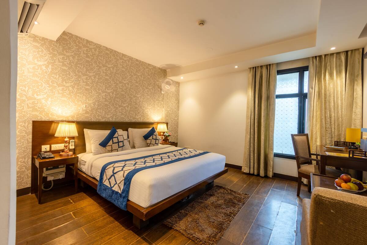 A hotel room with a large bed, a desk with a chair, a small table with a lamp, and a window with curtains.