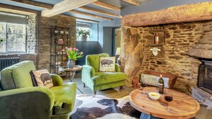 A cozy room with a stone wall, a fireplace, a round wooden table with a bottle and glass, and two green armchairs. - 4 Bed in Bridport (oc-dc212) (Burton Bradstock)