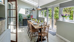 A dining area with a long table, chairs, pendant lights, and a view of greenery outside. - 4 Bed in Bridport (oc-dc212) (Burton Bradstock)