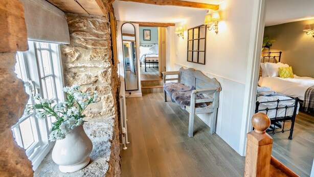 A rustic interior with a stone wall, wooden beams, a bench, a bed, and a hallway leading to another room. - 4 Bed in Bridport (oc-dc212) (Burton Bradstock)