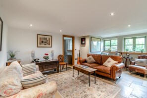 Ein Wohnzimmer mit braunem Ledersofa, einem Holztisch und einem weiteren Sofa mit Blumenmuster. Es gibt einen Esstisch mit Stühlen, eine Gitarre und einen Spiegel an der Wand.