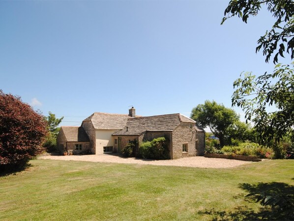 A stone house with a sloped roof, a front lawn, and surrounding vegetation. - 5 Bed in Swanage (oc-dc201) (Worth Matravers)
