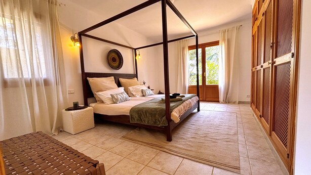 Una cama con dosel de madera, una mesita de noche, un banco tejido y vistas a una puerta de madera.
