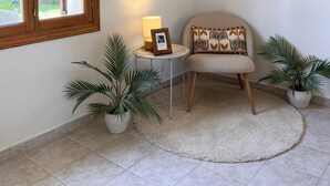 Una habitación con una silla, una pequeña mesa con una lámpara, dos plantas en macetas y una ventana.