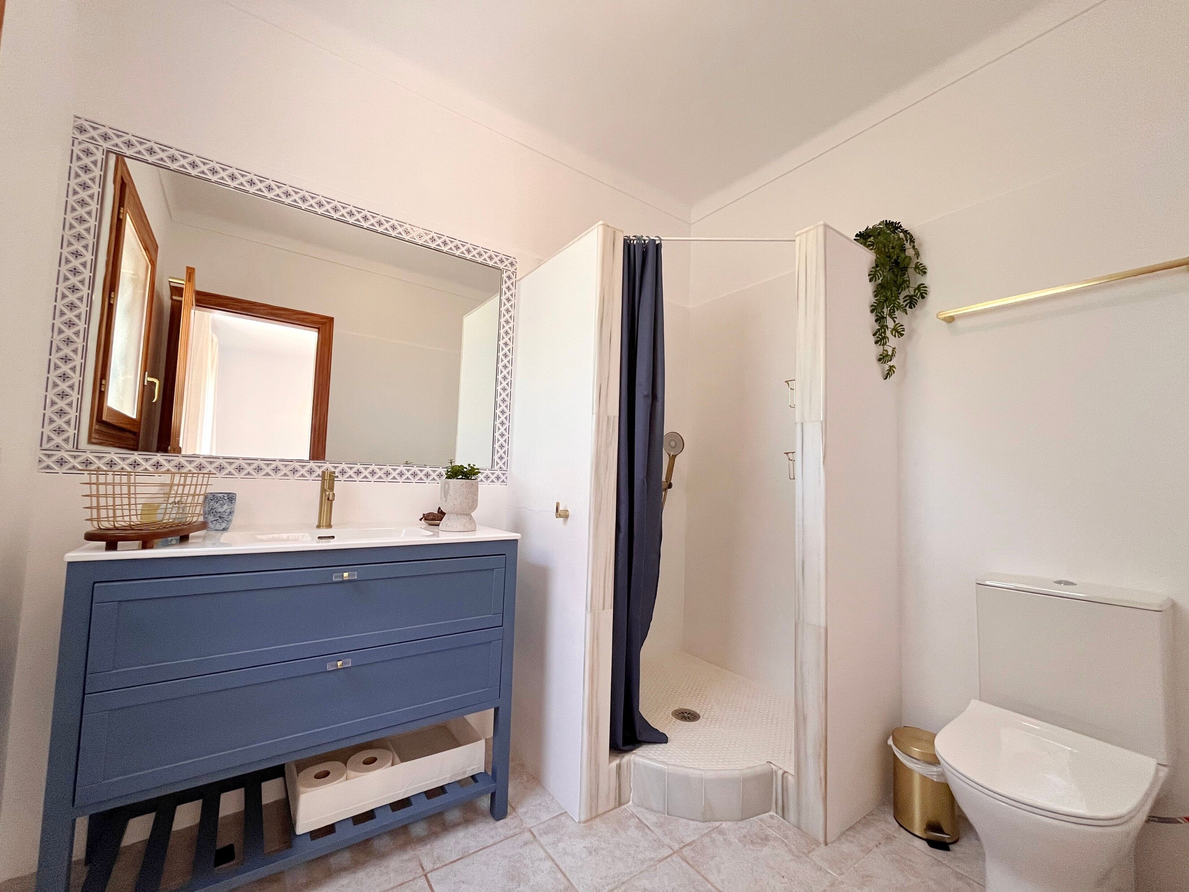 Baño con tocador azul, ducha, inodoro y espejo con detalles decorativos.