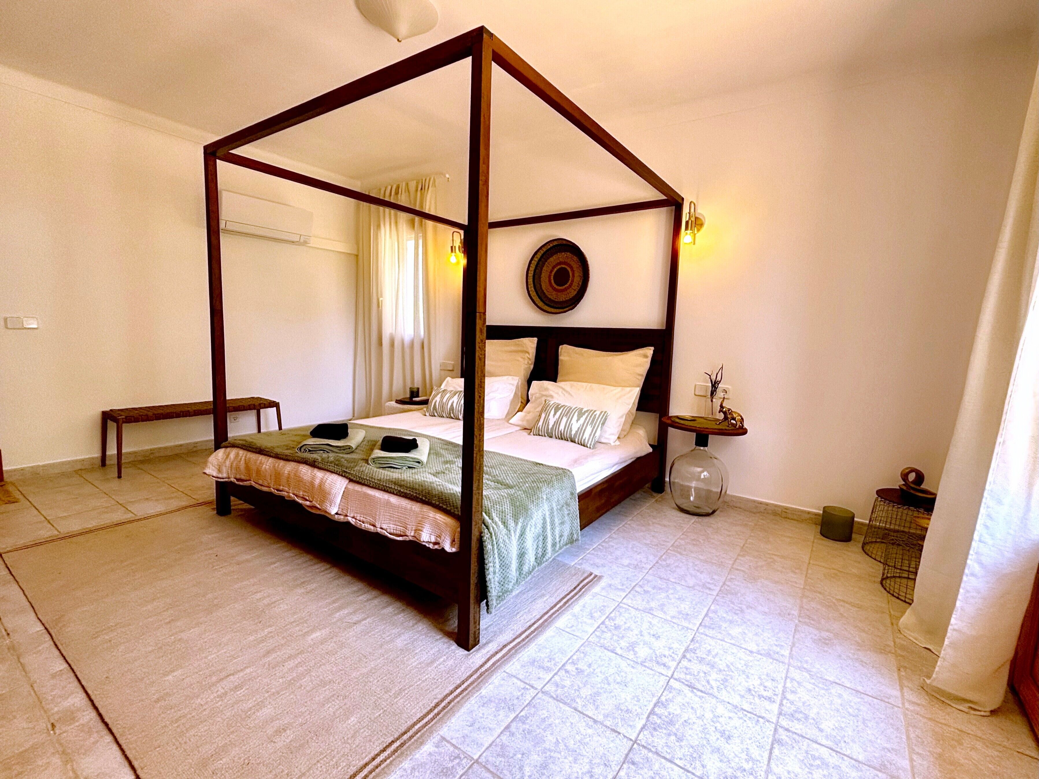 A four-poster bed in a room with a tiled floor, a bedside table with a lamp, and a small decorative item on the nightstand.