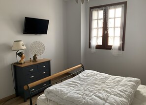 Un dormitorio con cama, cómoda con cajones, lámpara y televisión fijada a la pared.