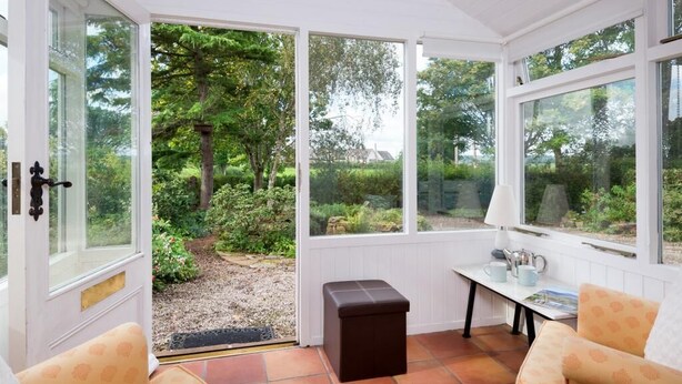 Ein Wintergarten mit einer Bank, einem Tisch mit Tasse und Untertasse sowie Blick auf einen Garten.