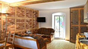 A room with a brick wall, wooden beams, a sofa, a chair, a TV, a door leading outside, and a kitchen area.