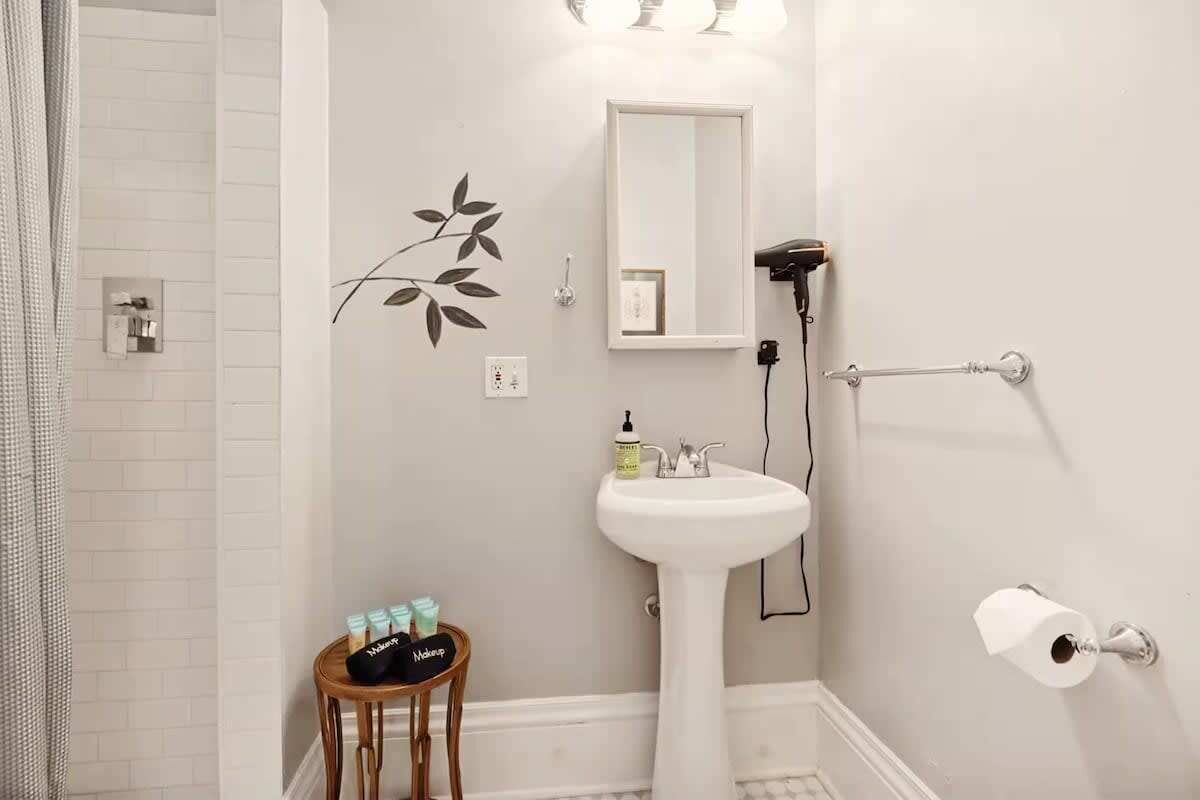 A bathroom with a white pedestal sink, a mirror, a towel rack, and a small stool with toiletries.