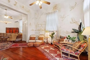 A living room with a wooden floor, a sofa, a chair, a small table, and a ceiling fan. - Relaxing Bohemian Villa w/ Heated Pool 5br 4ba (New Orleans)
