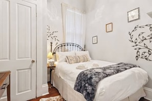 A bedroom with a bed, a nightstand, a lamp, and a window with curtains. - Relaxing Bohemian Villa w/ Heated Pool 5br 4ba (New Orleans)