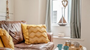 A brown leather sofa with a yellow pillow, a white table with blue cups and a plate of cookies, and a window with curtains and a decorative item. - 2 Bed in Brixham (oc-75550) (Brixham)