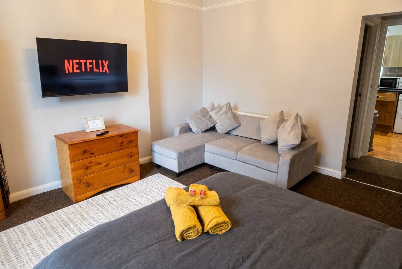 A hotel room with a bed, a grey sofa, a wooden dresser, a flat-screen TV, and a door leading to a bathroom.