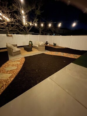 An outdoor seating area with a sofa, a chair, and a small table, surrounded by a white fence and string lights.