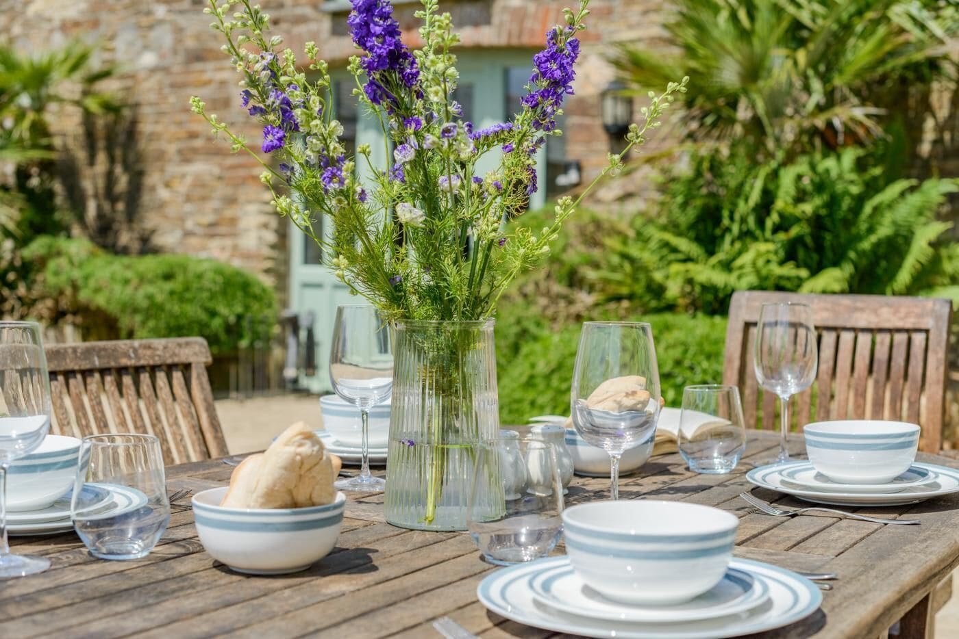 A wooden table set for dining with plates, bowls, glasses, and a vase with purple flowers.