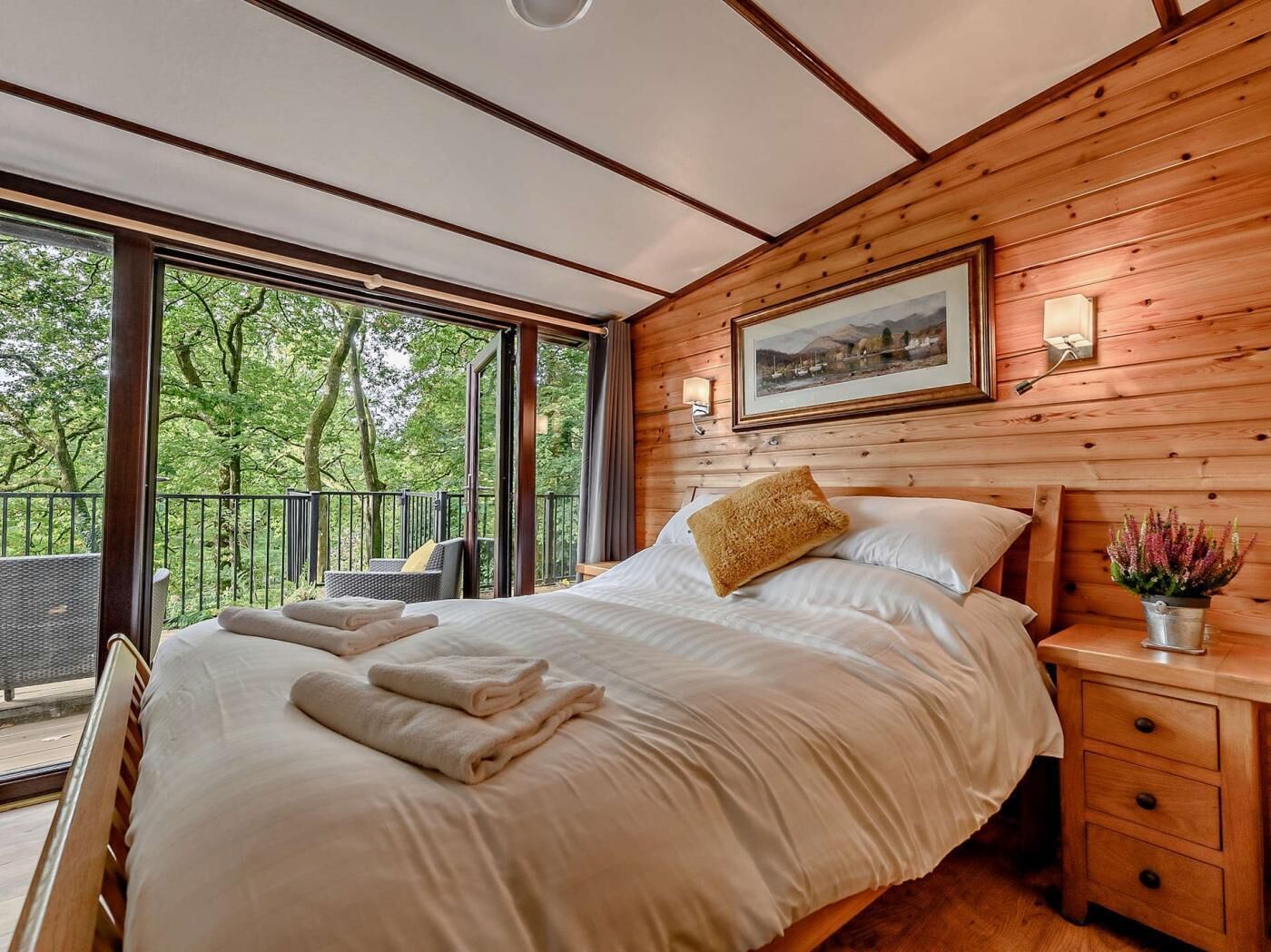 Ein Schlafzimmer mit einem großen Bett, einem Nachttisch, einem Fenster mit Blick auf Bäume und einem gerahmten Bild an der Wand.