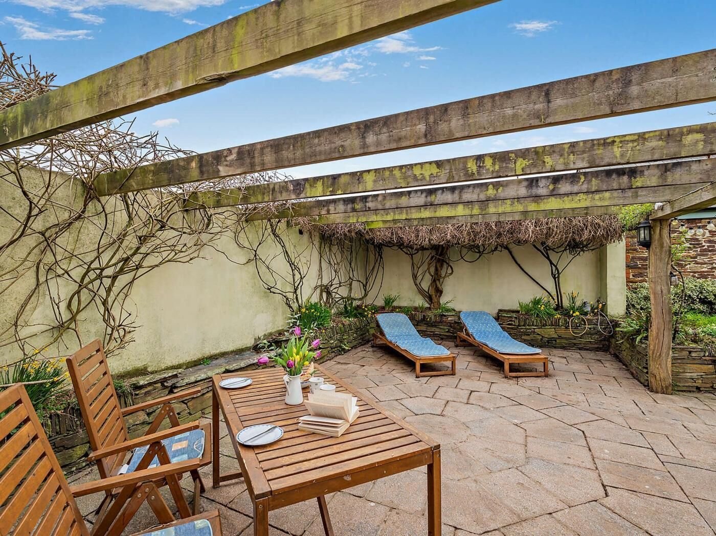A patio with wooden furniture, a table set for two, and two lounge chairs under a pergola.