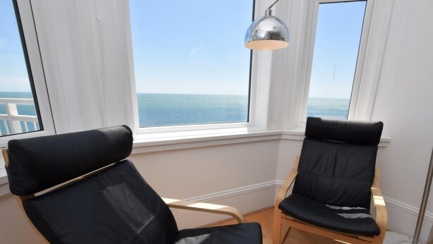 A room with two black cushioned chairs, a wooden floor, a large window with a view of the sea, and a floor lamp. - 2 Bed in Ventnor (oc-75730) (Ventnor)
