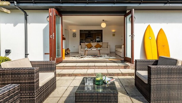 Eine Terrasse mit Korbmöbeln, einem Glastisch und zwei Surfbrettern, die an der Wand lehnen.