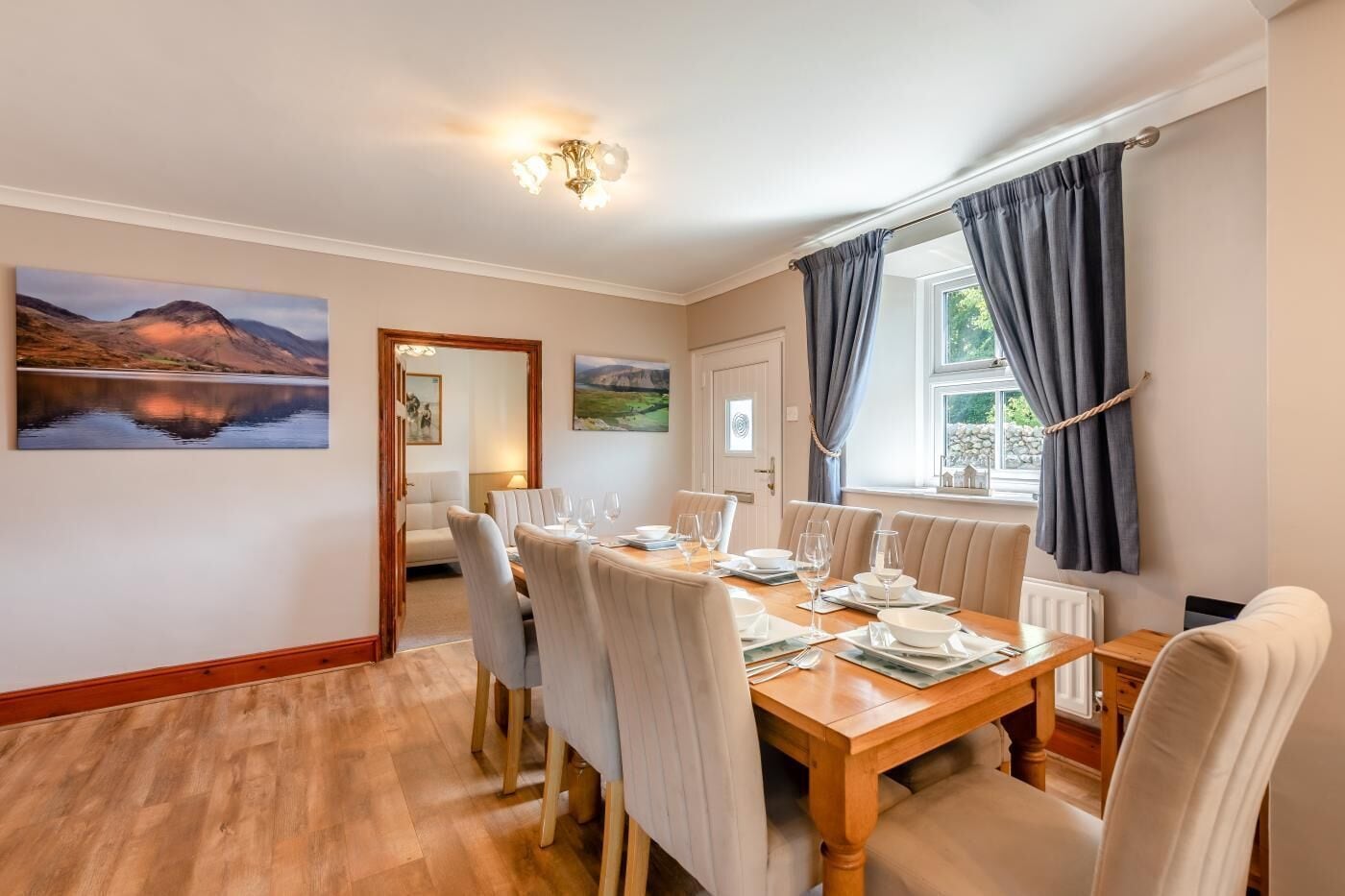 A dining area with a wooden table set for four, surrounded by chairs, with a painting of a landscape on the wall.