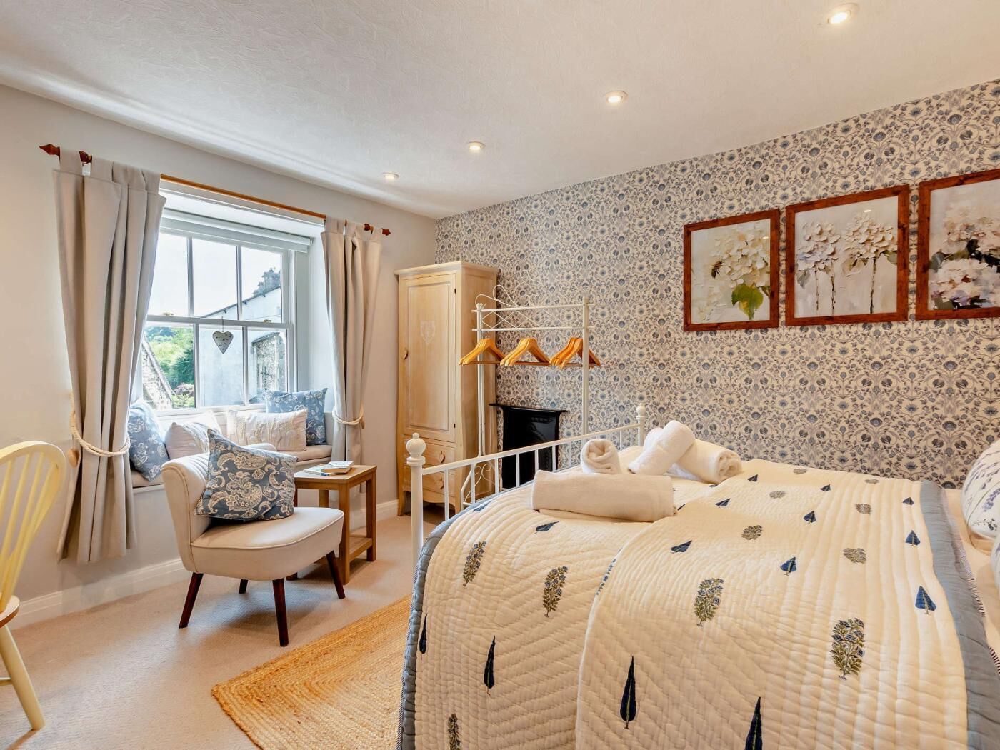 A bedroom with a bed, a chair, a wardrobe, a window with curtains, and a wall with framed pictures.