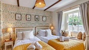 A hotel room with two beds, floral wallpaper, framed botanical prints, a window with curtains, and a hanging light fixture. - 3 Bed in Ulverston (oc-sz445) (Satterthwaite)