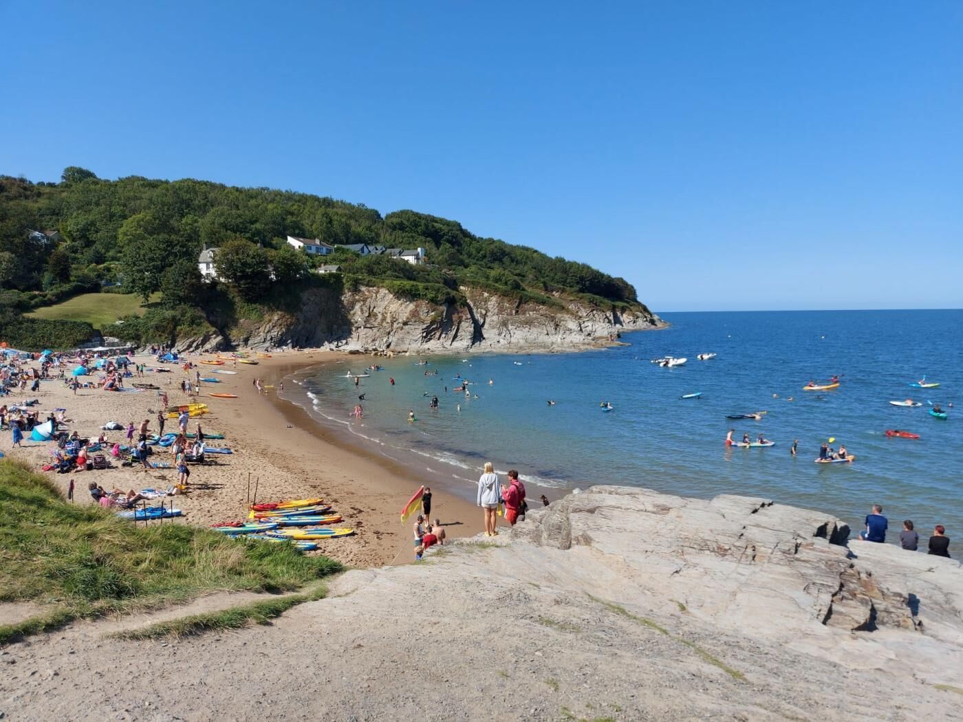 A beach with kayaks and people, a rocky shoreline, and houses on a hill.