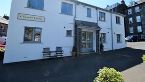 A two-story building with a sign reading 'Chaucer Lodge.' There are two windows on the ground floor, a door with a stone facade, and a small porch area with two benches. - 1 Bed in Keswick (oc-85419) (Keswick)