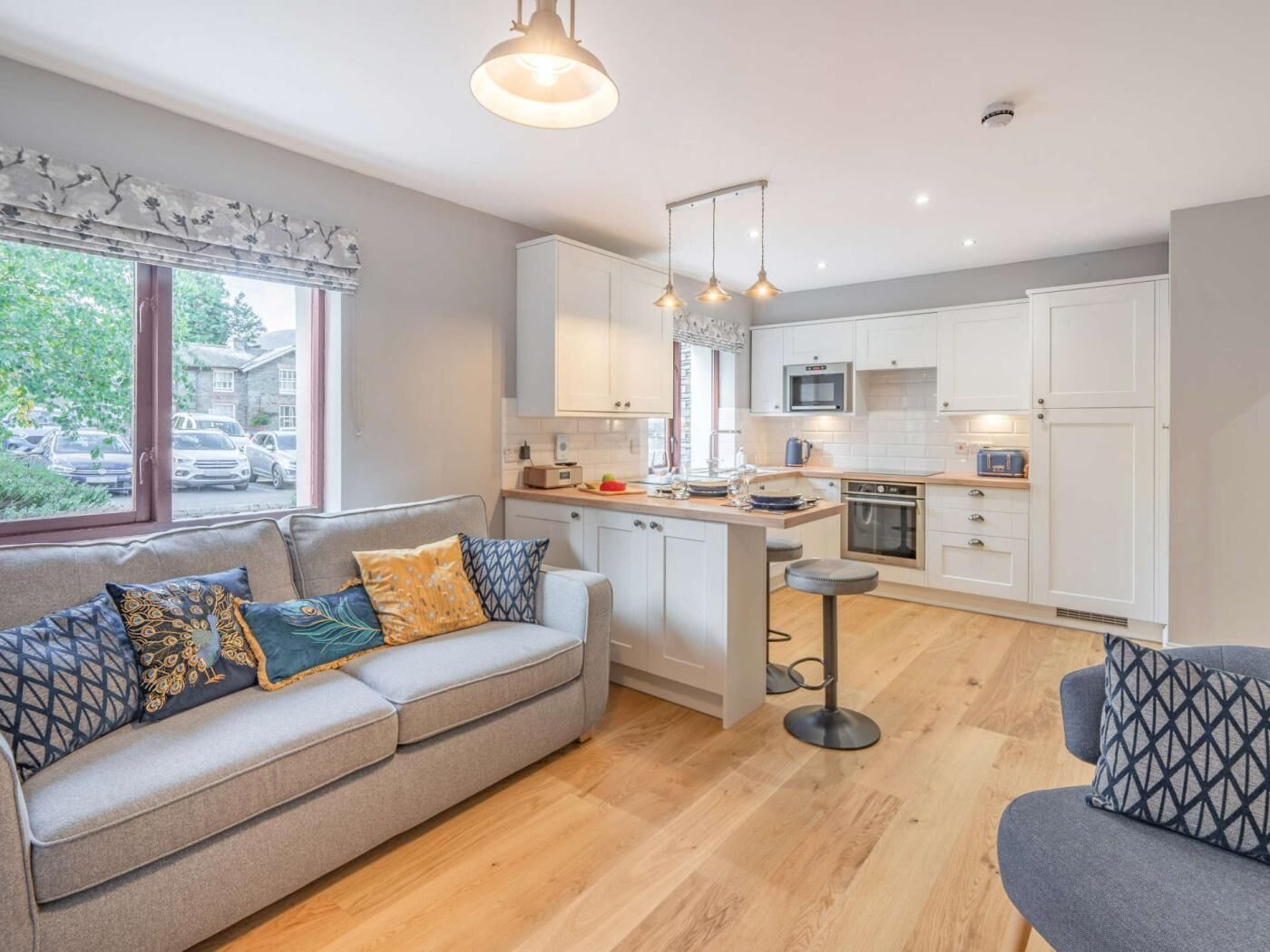 A modern kitchen with white cabinets, a central island, and a dining area with a grey sofa and patterned cushions.