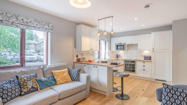 A modern kitchen with white cabinets, a central island, and a dining area with a grey sofa and patterned cushions. - 1 Bed in Keswick (oc-85419) (Keswick)
