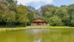A wooden cabin with a porch, surrounded by a pond and dense greenery. - 1 Bed in Holsworthy (oc-85561) (Bradworthy)
