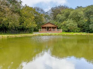 A wooden cabin with a porch, surrounded by a pond and dense greenery. - 1 Bed in Holsworthy (oc-85561) (Bradworthy)