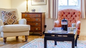 Ein gemütliches Zimmer mit Holzboden, einem weißen Sessel, einem gemusterten Teppich, einem hölzernen Kommode, einem pinken, gepolsterten Sessel, einem Couchtisch mit Kaffeekanne und gerahmten Kunstwerken an der Wand.