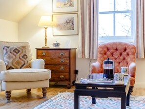 A cozy room with a wooden floor, a white armchair, a patterned rug, a wooden dresser, a pink tufted armchair, a coffee table with a coffee pot, and framed artwork on the wall. - 3 Bed in Achnasheen (oc-ca120) (Inveralligin)