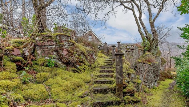 Eine moosbewachsene Steinsteintreppe führt zu einem traditionellen Steinhaus, umgeben von Bäumen.