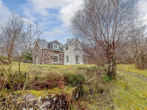 A stone house with a gabled roof, surrounded by a natural landscape with trees and a grassy area. - 3 Bed in Achnasheen (oc-ca120) (Inveralligin)