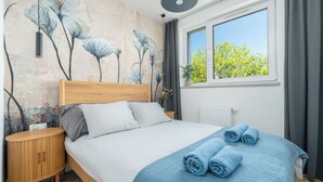 A bedroom with a wooden bed, a nightstand, a wall-mounted lamp, a window with curtains, and a floral mural. - SeaLight Apartment – Cozy Apartment in the Heart of Świnoujście (Swinemünde)