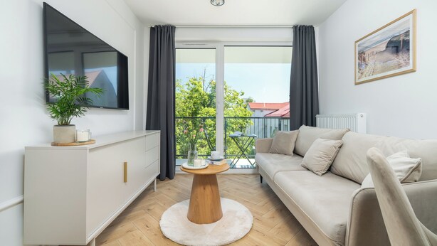 A modern living room with a sofa, a round wooden coffee table, a TV on the wall, and a balcony with a view of greenery. - SeaLight Apartment – Cozy Apartment in the Heart of Świnoujście (Swinemünde)