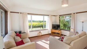 Ein modernes Wohnzimmer mit Fernseher, zwei Sofas, einem kleinen Tisch und großen Fenstern mit Blick auf Grünflächen.