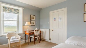 Une chambre à coucher avec un lit, une chaise, un bureau avec une lampe, une fenêtre donnant sur un paysage agréable et une porte.