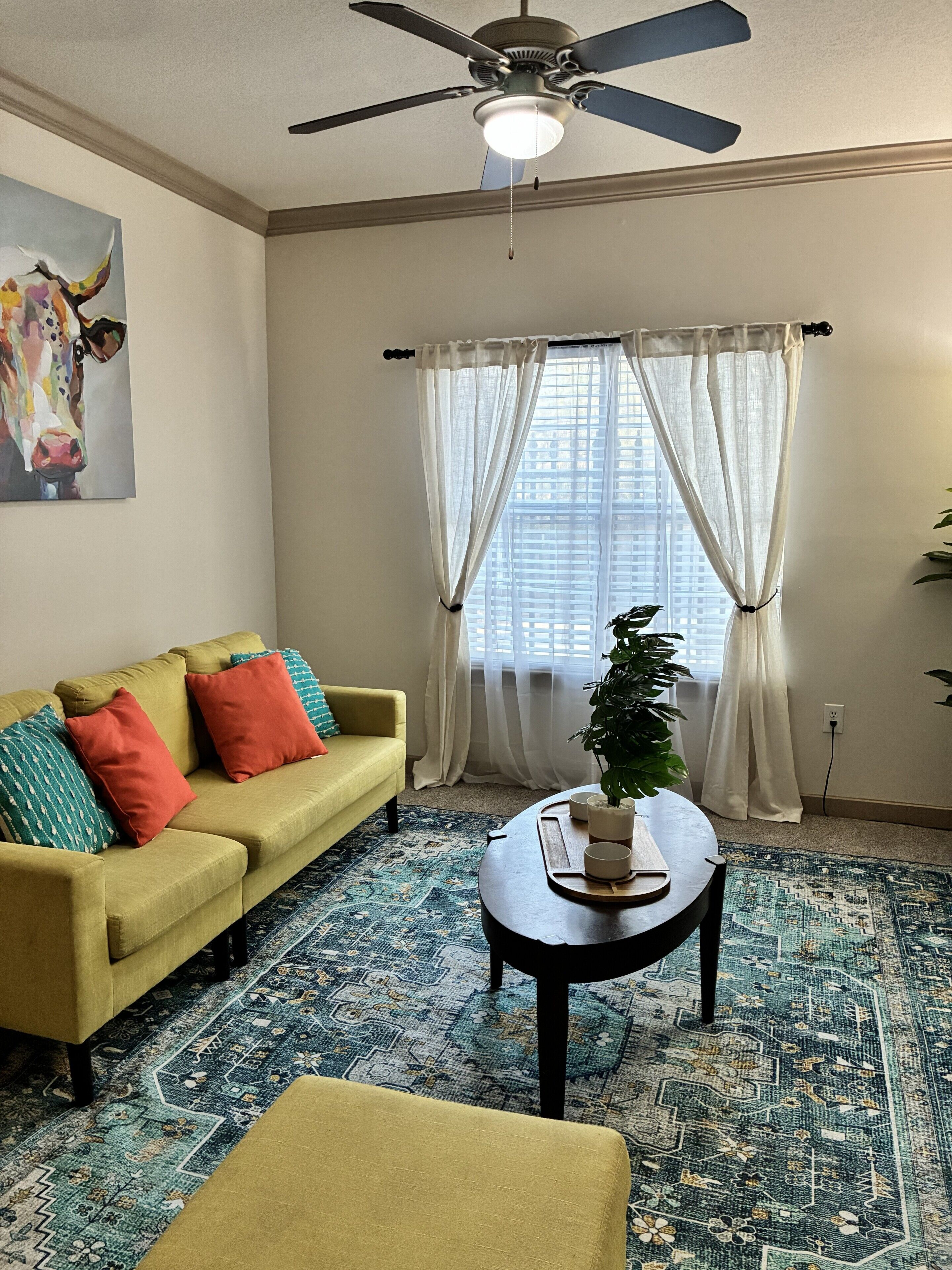 A living room with a yellow sofa, a coffee table, a ceiling fan, and a window with sheer curtains.